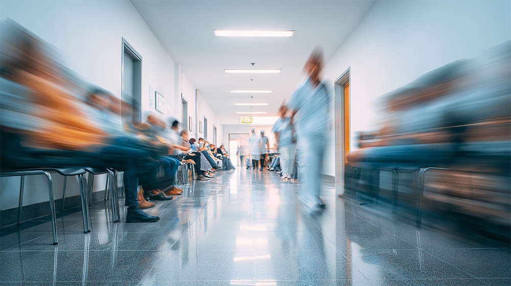 A long, bright hospital corridor with rows of seated patients on both sides and medical staff walking through the middle, all shown with motion blur to suggest a busy, crowded waiting area.