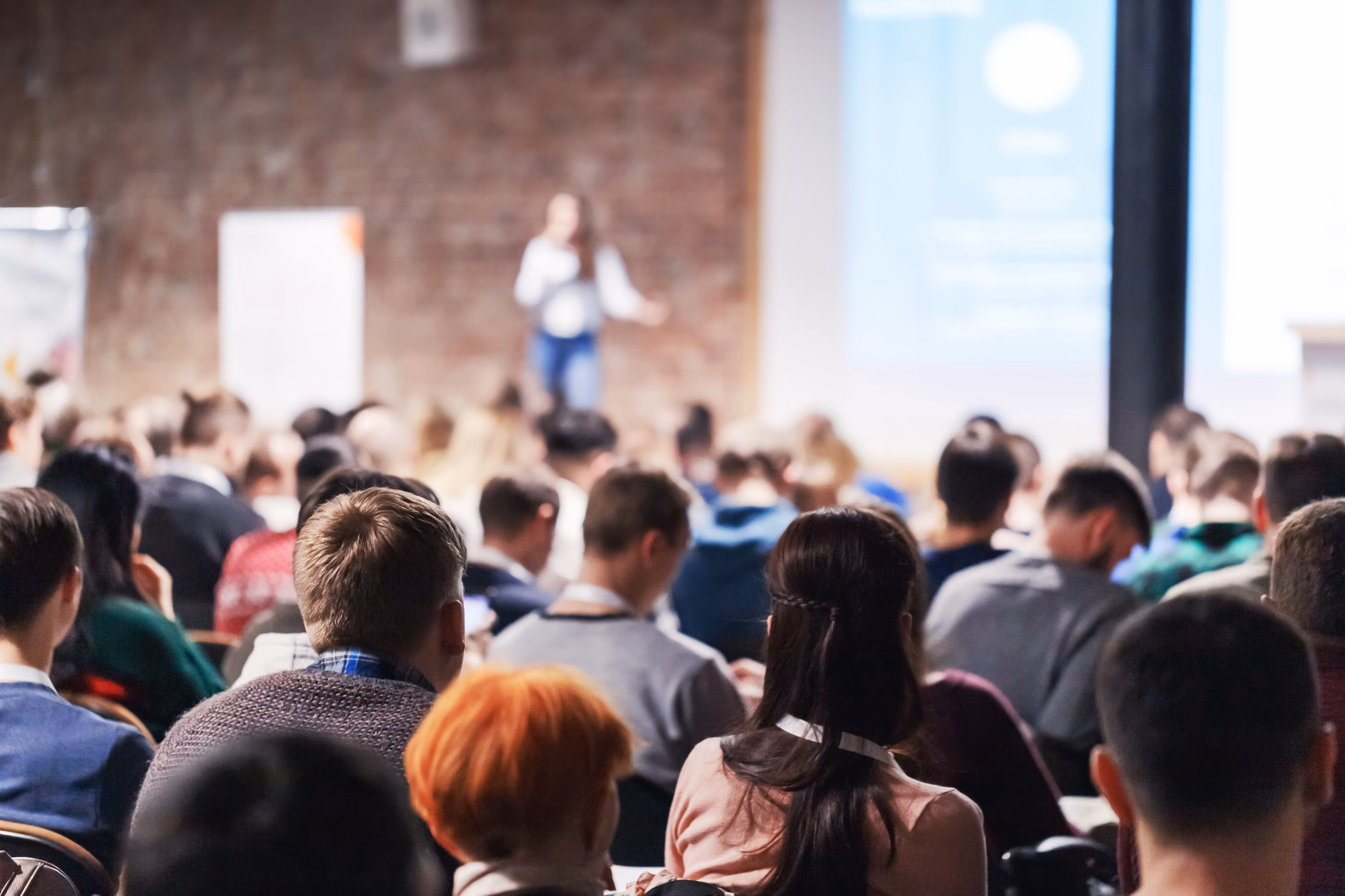 Audience seated and listening to a speaker at a conference or seminar in a large room.