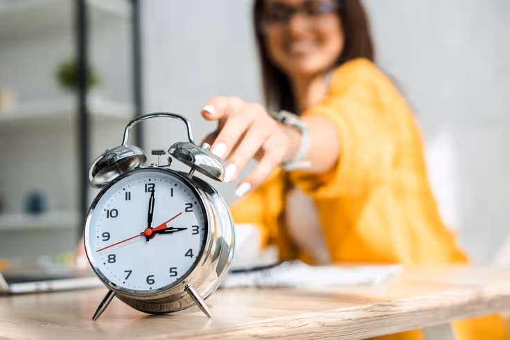 Silver analog alarm clock showing 3:00 on a wooden table with a blurred person in a yellow shirt reaching for it.