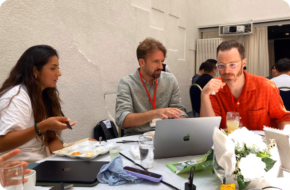 Three people seated at a table engaged in discussion with an open MacBook laptop, drinks, and a plate with food on the table.
