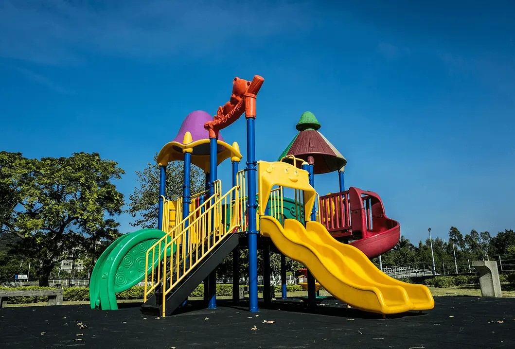 Colorful outdoor playground with yellow double slides, green curved slide, stairs, and covered climbing structures against a clear blue sky.