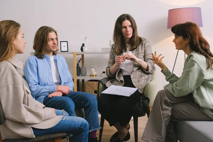 Three individuals sitting in a therapy session in a cozy room with a lamp and shelves.