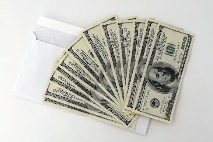 A spread of US $100 bills fanned out from an envelope on a white background.