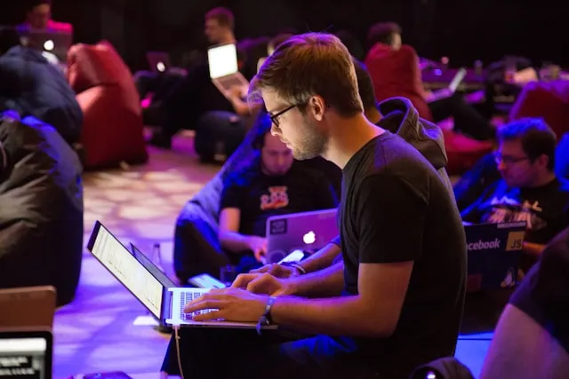 Man using a laptop at a tech event with other attendees working in the background.