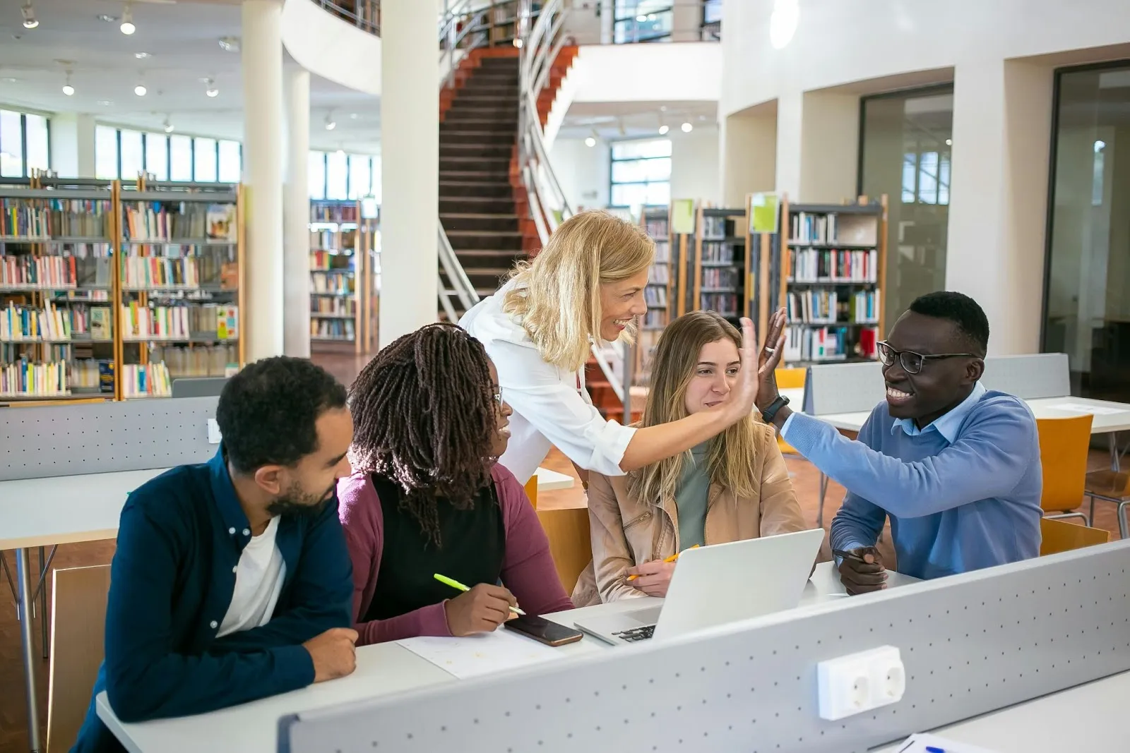 Group of diverse people collaborating at a table in a library with books and staircase.