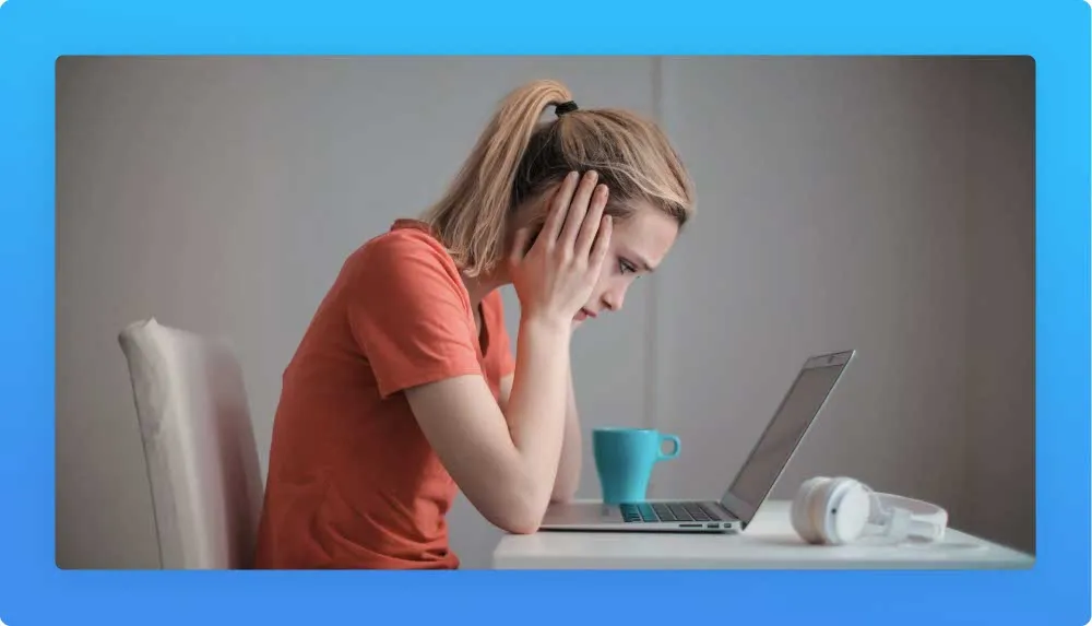 Profile of a young woman at her desk showing frustration signals while looking at the laptop. Blue mug and white headphones on the desk.  