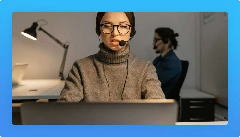 Young administrative woman with headphones and mic during a call. Co-worker in the the background