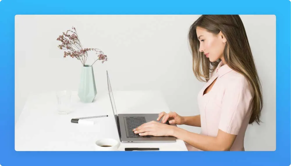 Left profile of seated woman in a pink suit with hands on laptop keyboard. On the desk also pens, cup and jar with wildflowers