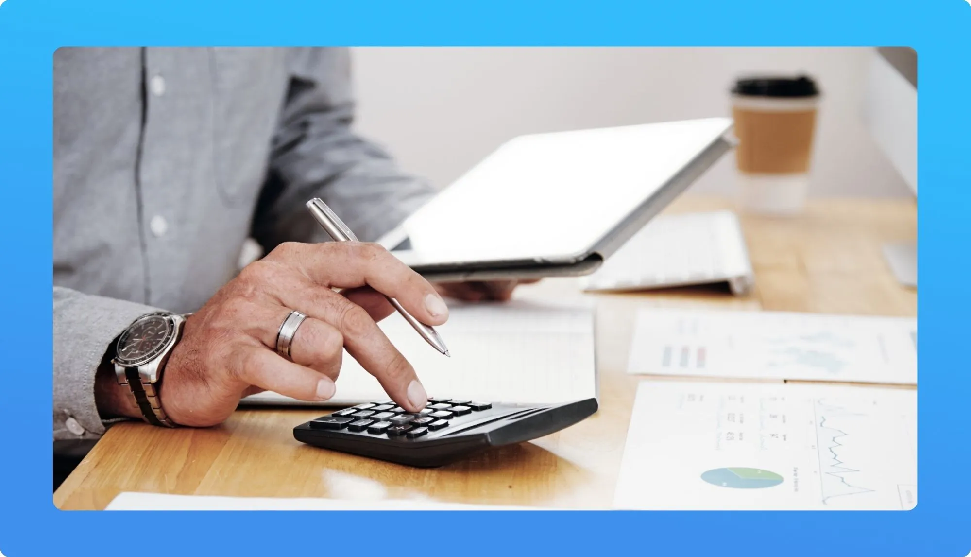 A person calculates numbers using a calculator while holding a tablet and jotting notes, surrounded by charts and a coffee cup.