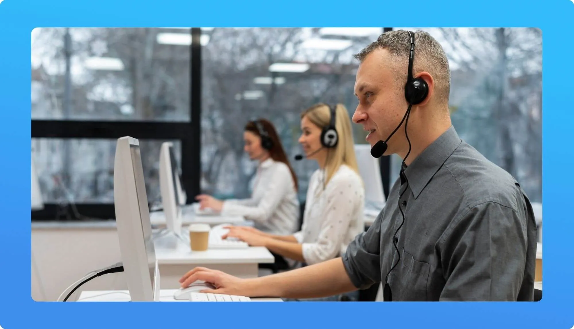 A man in a grey shirt sits at a computer, using a headset, with two women working at nearby desks in a modern office setting.