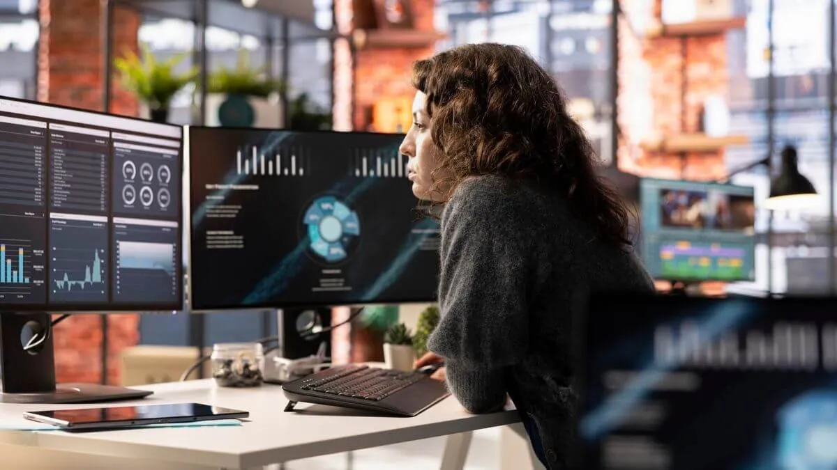 A person sits at a modern desk, focused on multiple monitors with data visualizations and analytics, surrounded by greenery.