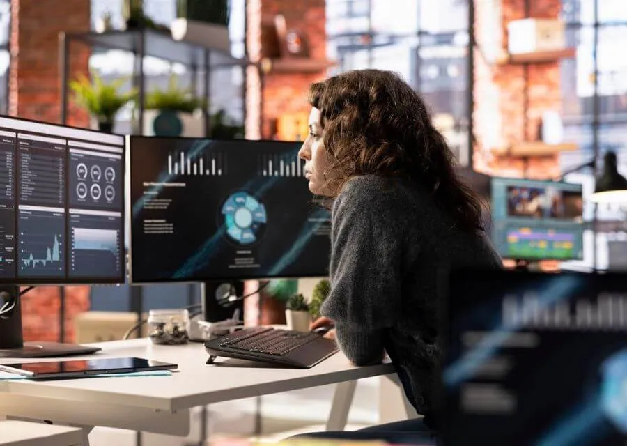 A person sits at a modern desk, focused on multiple monitors with data visualizations and analytics, surrounded by greenery.