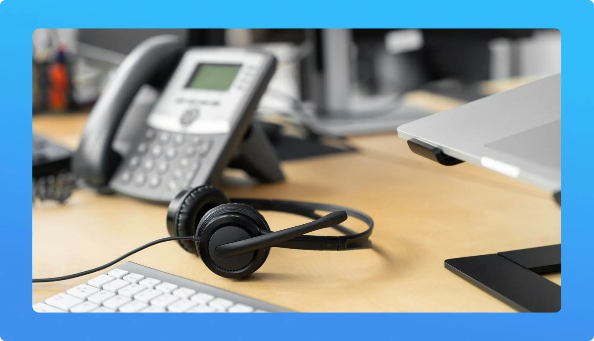 A black headset rests on a wooden desk beside a keyboard and a digital phone, with a laptop stand in the background.