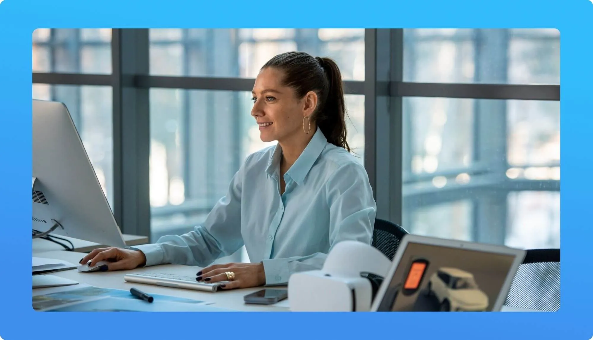 A woman in a light blue blouse works at a desk with a computer, beside a tablet and a smartphone, in a bright office space.