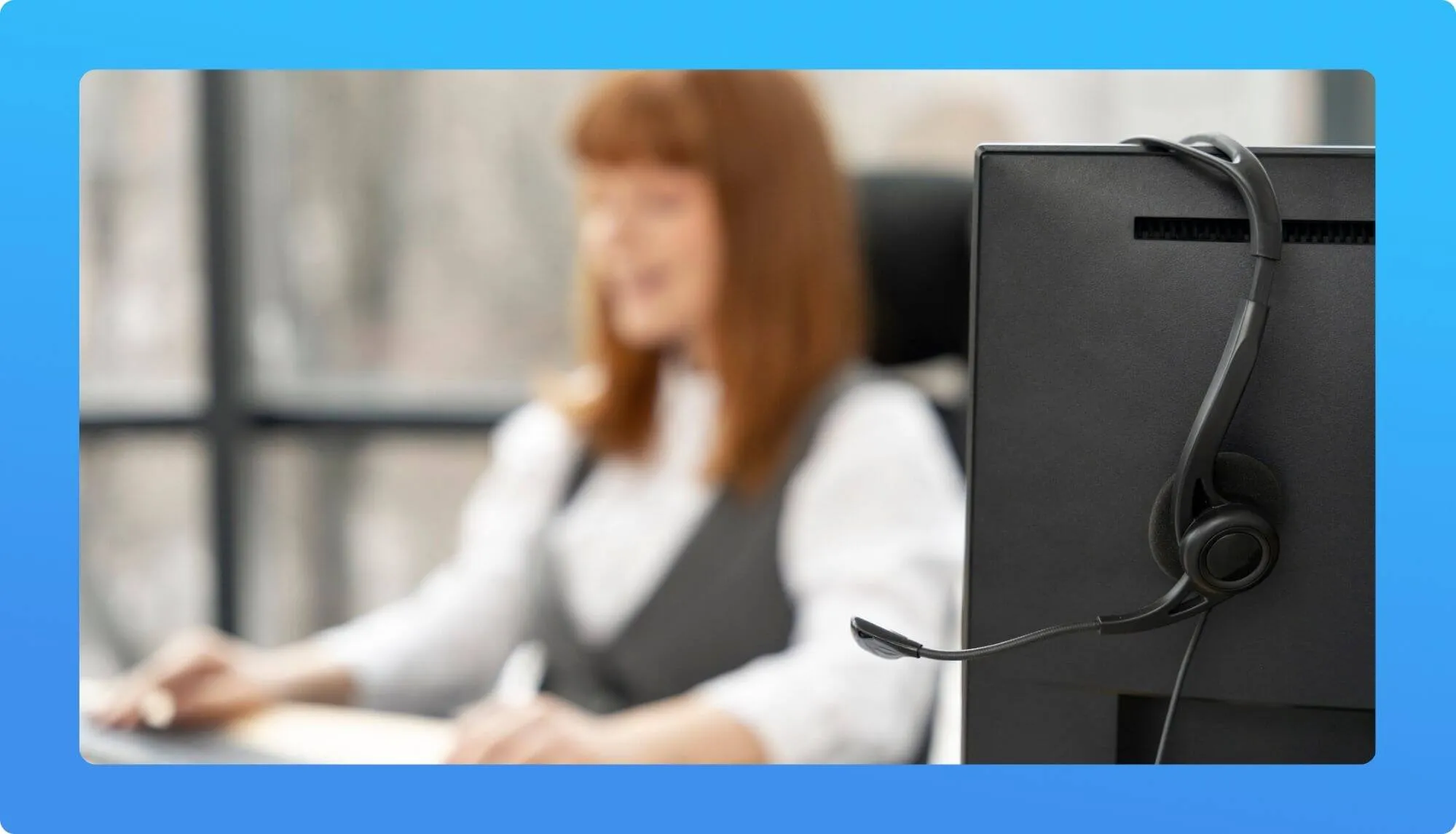A headset rests on the back of a computer monitor, with a blurred office scene and a person working on a desk in the foreground.