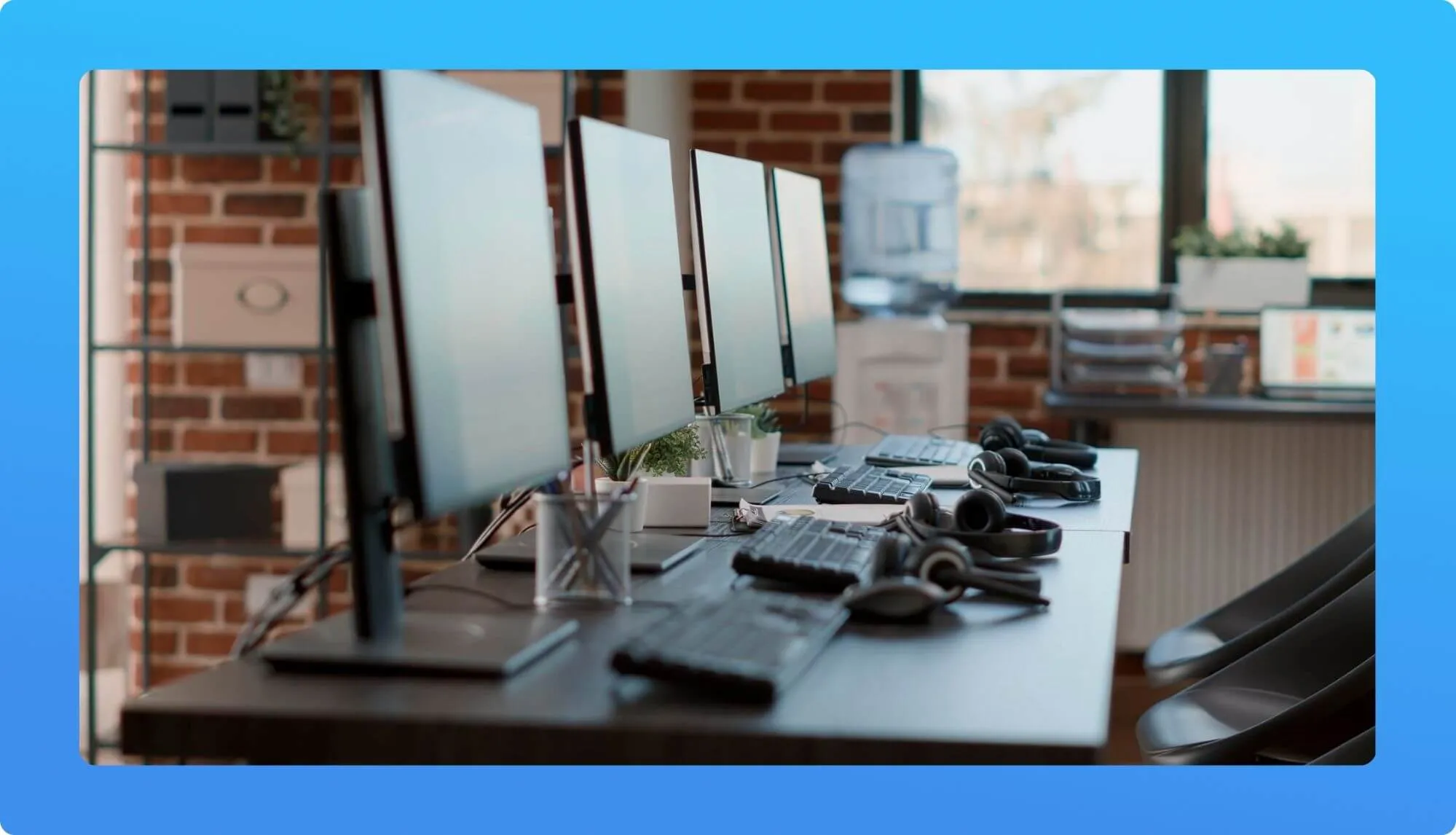 A modern office workspace featuring multiple monitors, keyboards, headphones, and a water cooler against a brick wall backdrop.