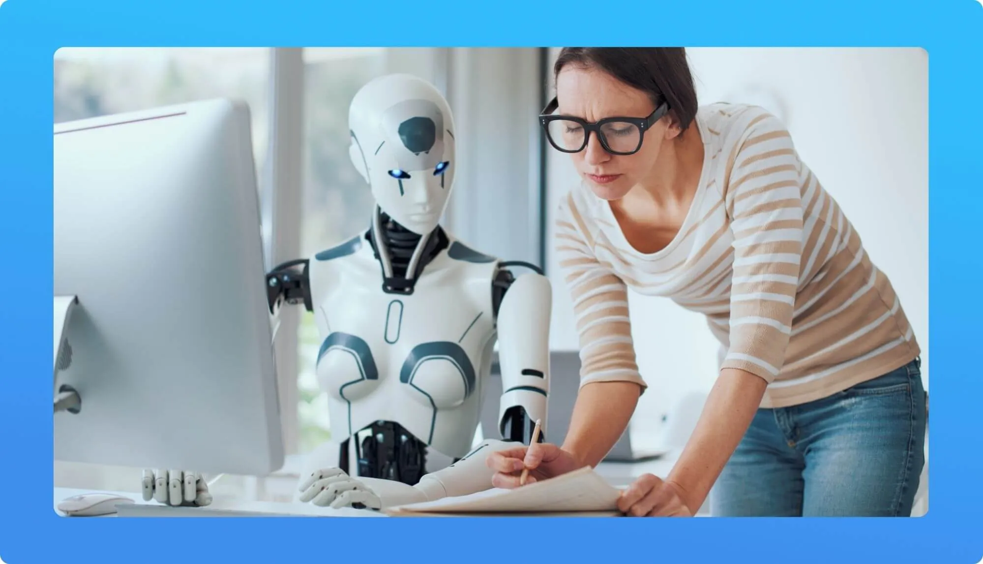 A person in a striped shirt writes notes beside a humanoid robot, with a computer monitor visible in a bright, modern workspace.