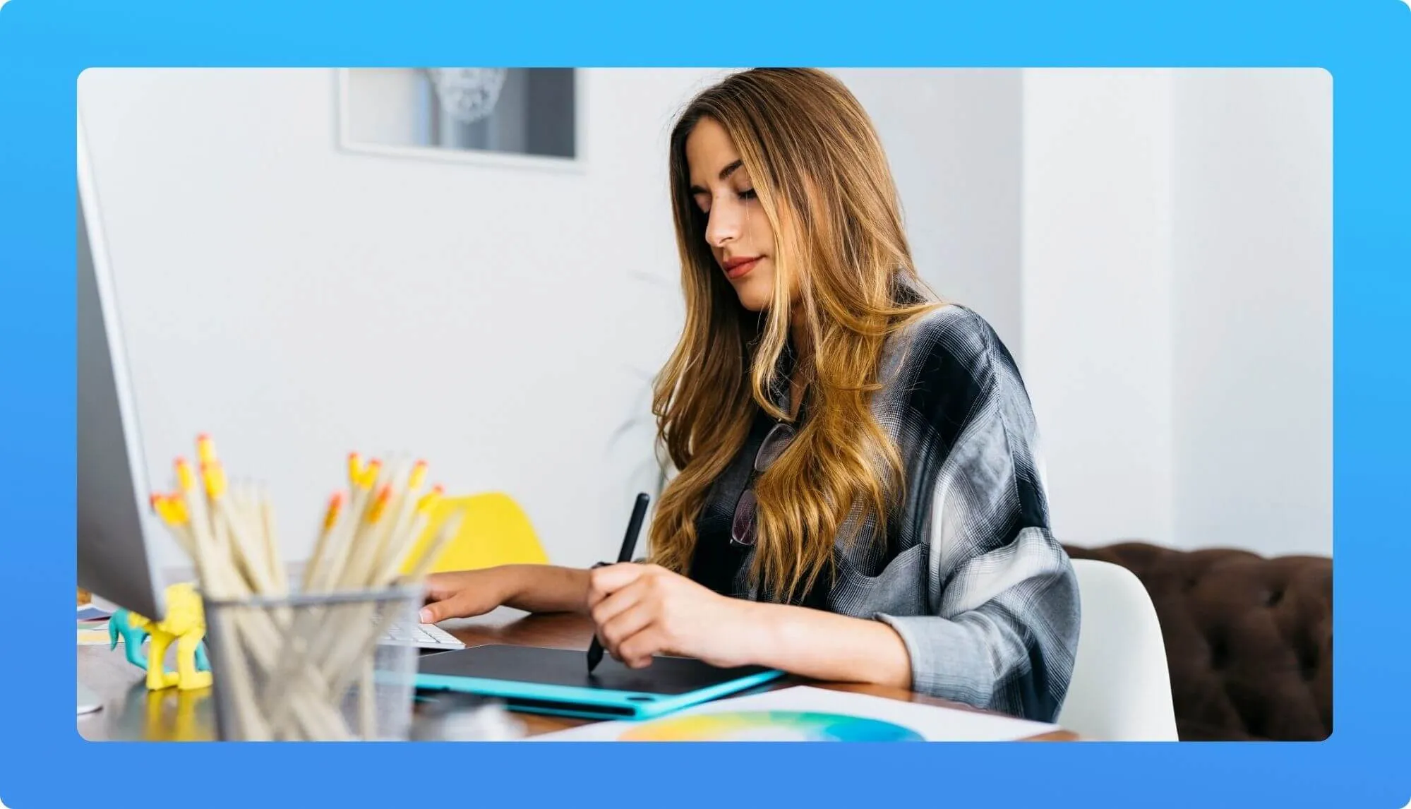 A person with long hair wearing a patterned shirt sits at a desk, working on a laptop surrounded by colorful stationery and office decor.