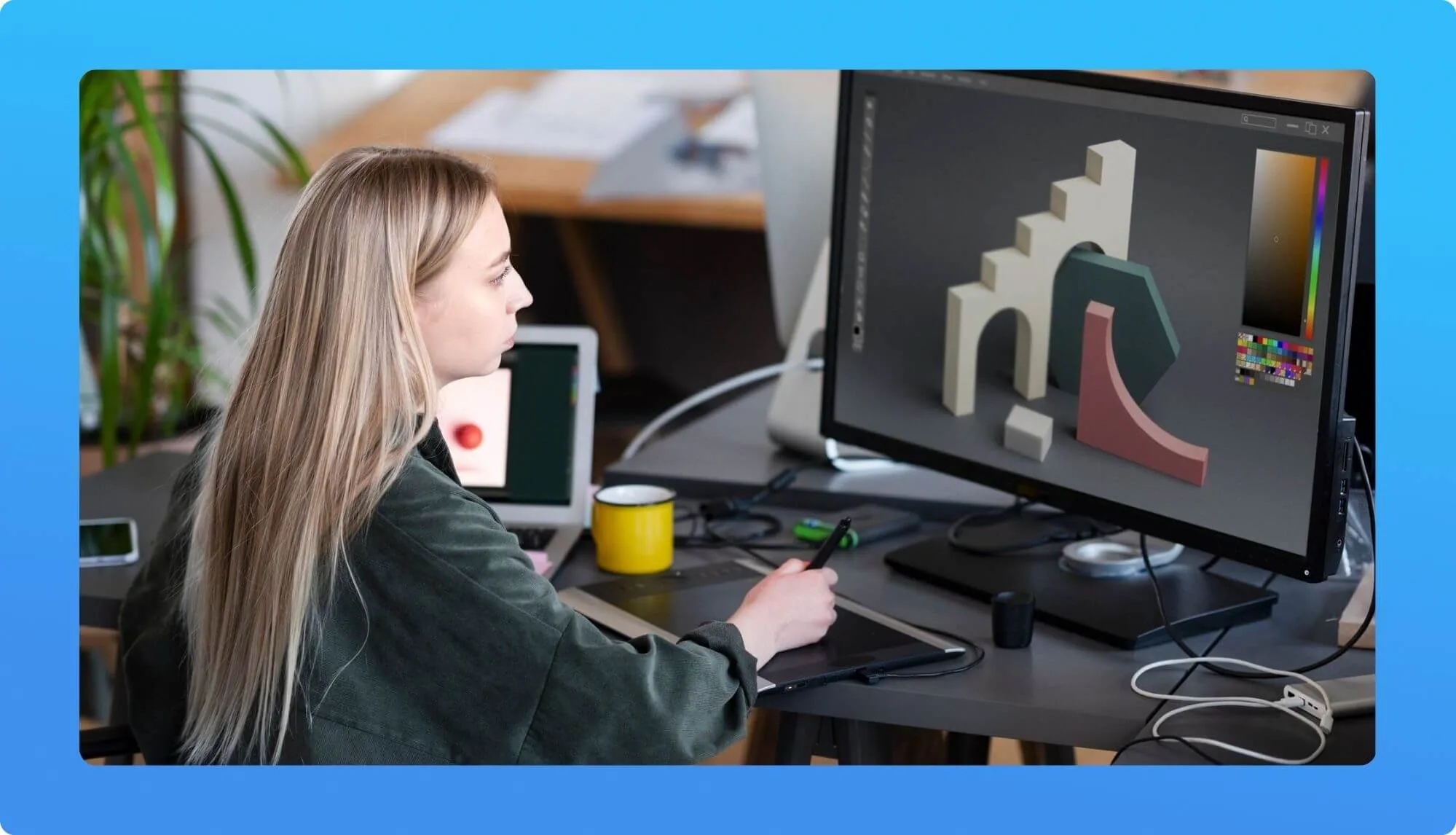 A person works on a digital tablet at a desk, surrounded by a laptop, a yellow mug, and a vibrant computer display of 3D shapes.