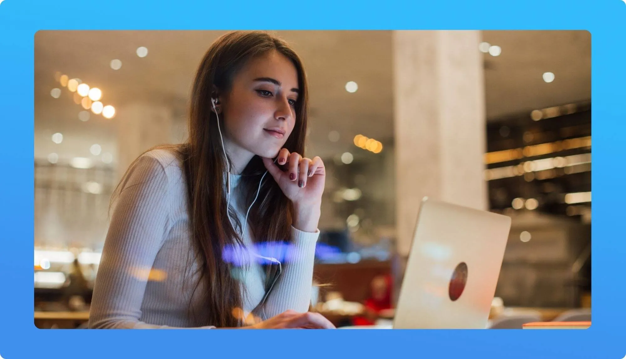 A young woman sits in a stylish café, focused on her laptop while wearing earphones, surrounded by soft, ambient lighting.