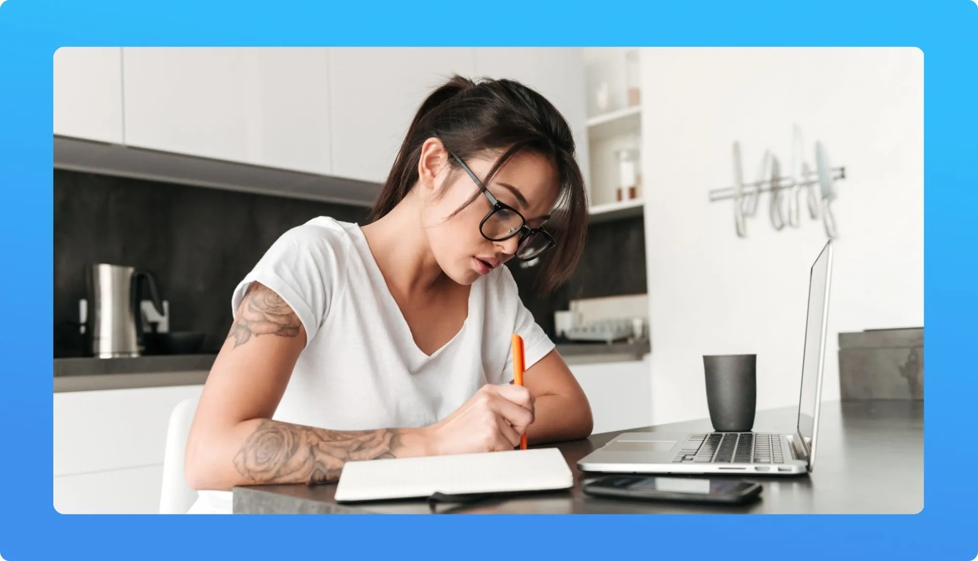A person sits at a wooden table writing in a notebook, with a laptop and a black mug nearby in a modern kitchen setting.
