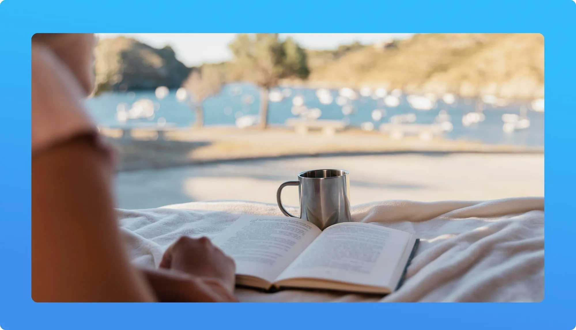 A person relaxes with a book and a metal mug of coffee beside a serene lake view, framed by soft blankets.