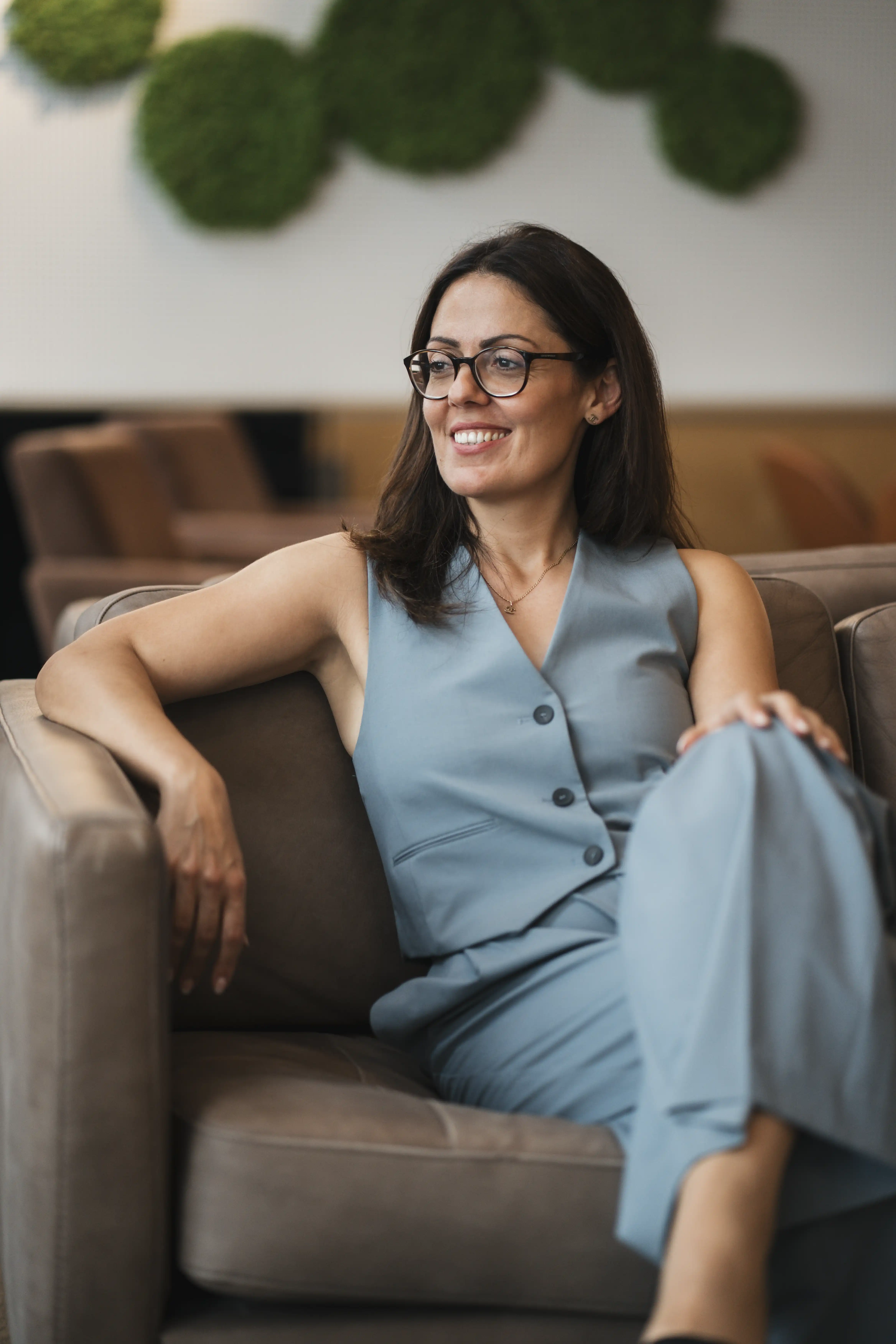 Anna Danieljan mit Brille, bekleidet mit einer blauen, ärmellosen Weste und einer Hose, sitzt auf einem braunen Sofa in einem modernen Raum.