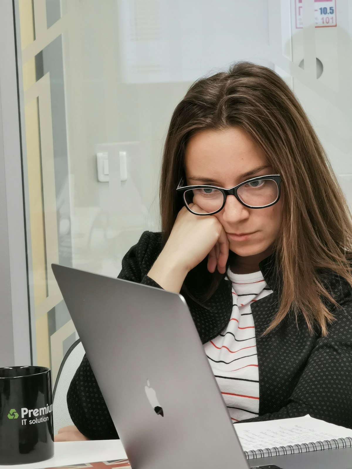Eine Frau mit Brille sitzt im Büro am Laptop, das Kinn in die Hand gestützt, und denkt nach.