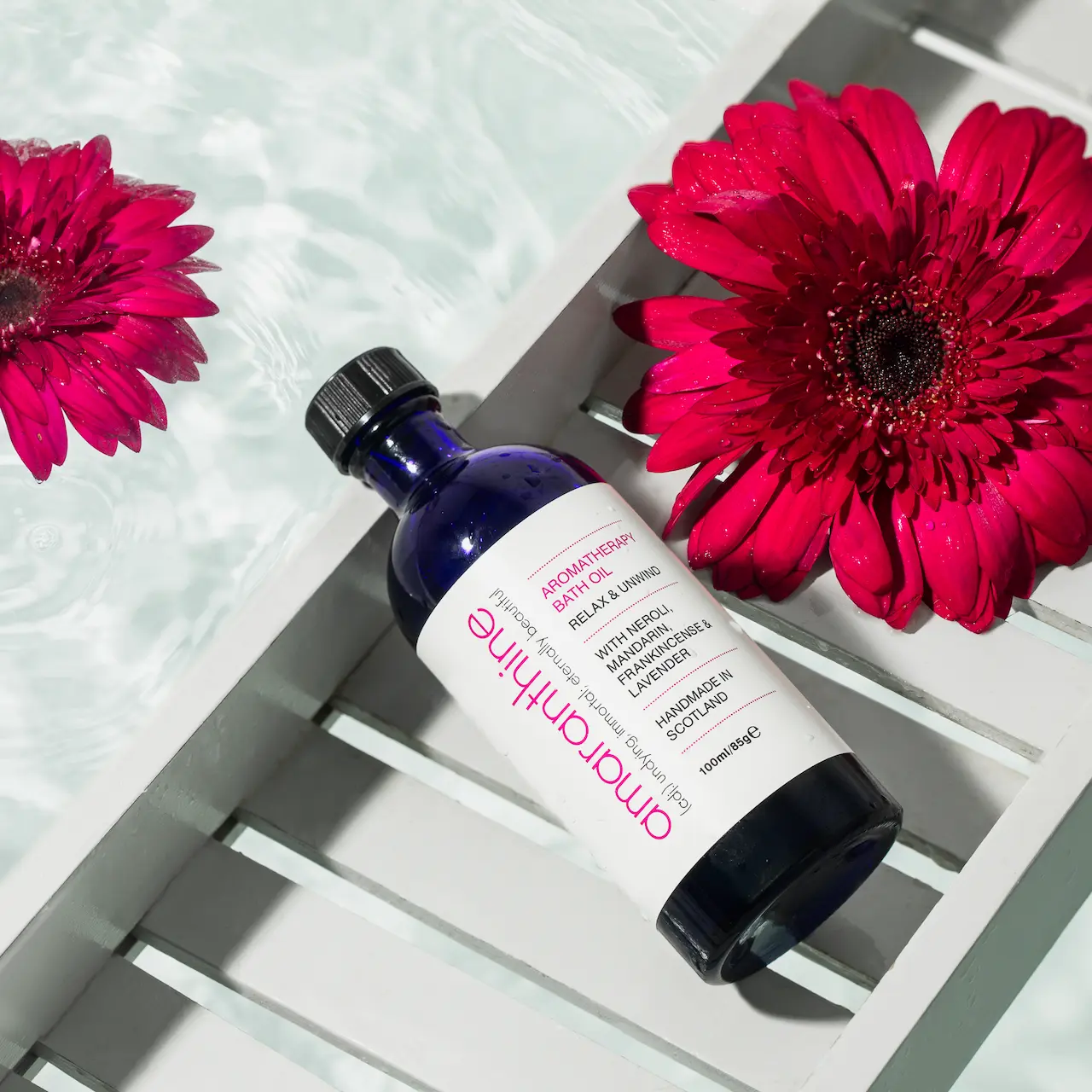 Bath oil on bath rack, above a filled bath with flowers, the perfect self care gift.