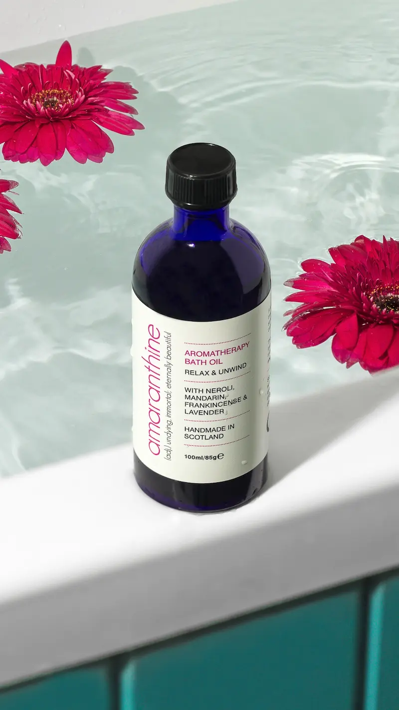 Amaranthine aromatherapy bath oil in a blue glass bottle resting on the edge of a bath, with pink flowers floating in milky bath water.