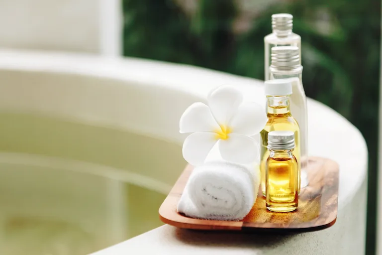 Aromatherapy bath oils resting on the edge of a bath, sitting on a wooden tray with a wash cloth and a white flower.
