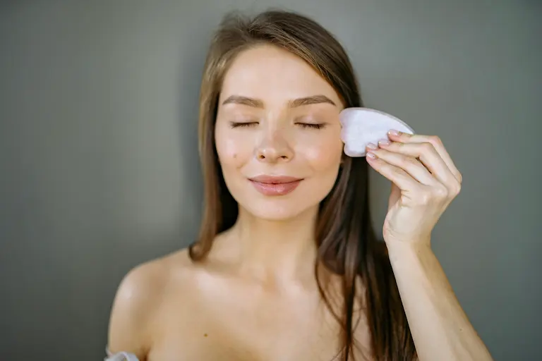 Woman performing a gua sha face massage with a white stone tool on smooth, well-oiled skin, demonstrating gentle facial massage technique.