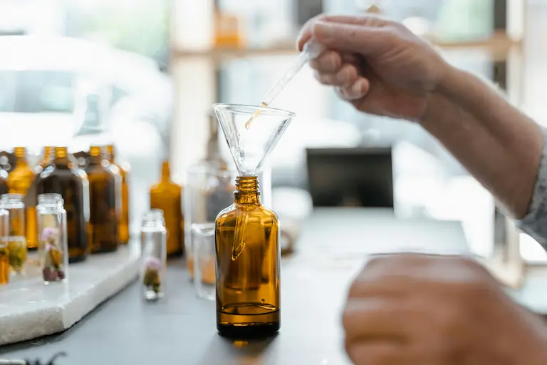 A person using a dropper to blend essential oil into a glass bottle through a funnel, surrounded by amber bottles and lab tools.