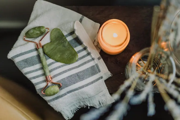 A candle, jade roller, and gua sha tool arranged on a striped towel, evoking a calming skincare ritual.
