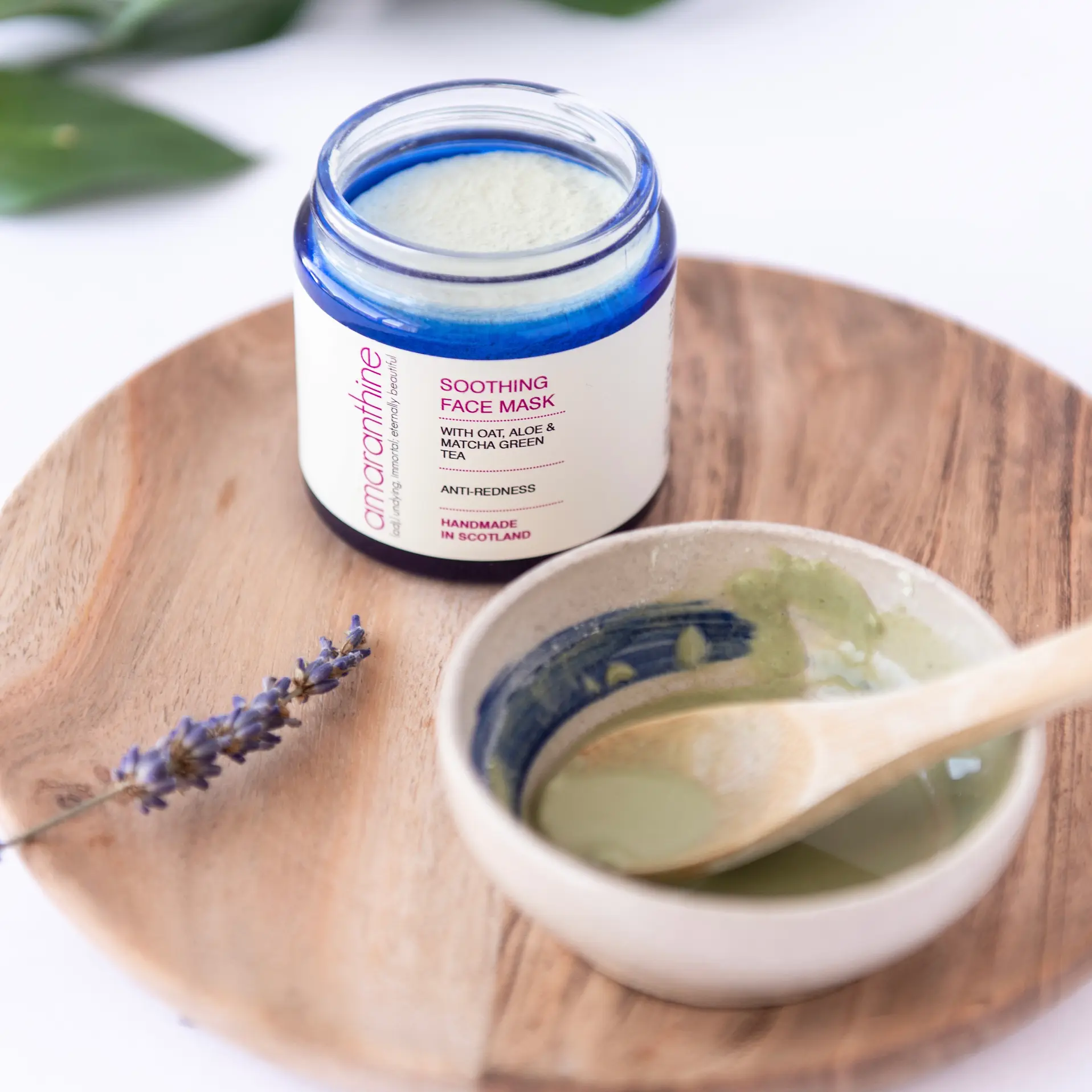 Soothing face mask for rosacea in blue glass jar with oat and matcha clay mixture in bowl on wooden tray.