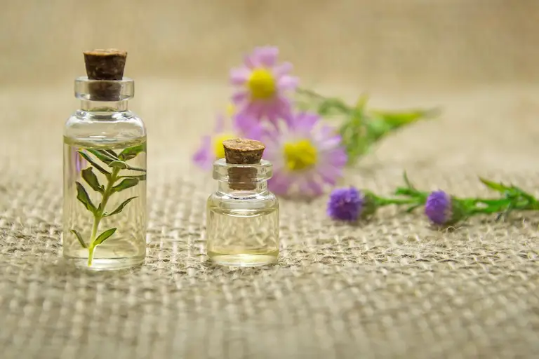 Small glass bottles of natural essential oils with botanical flowers on a neutral fabric background, representing calming aromatherapy ingredients.