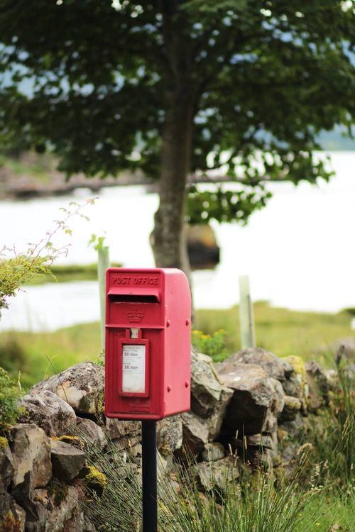A red Royal Mail post box on a rural road with a stone wall and fields and trees in the background, depicting that Amaranthine uses Royal Mail for delivering their natural beauty and skincare products.
