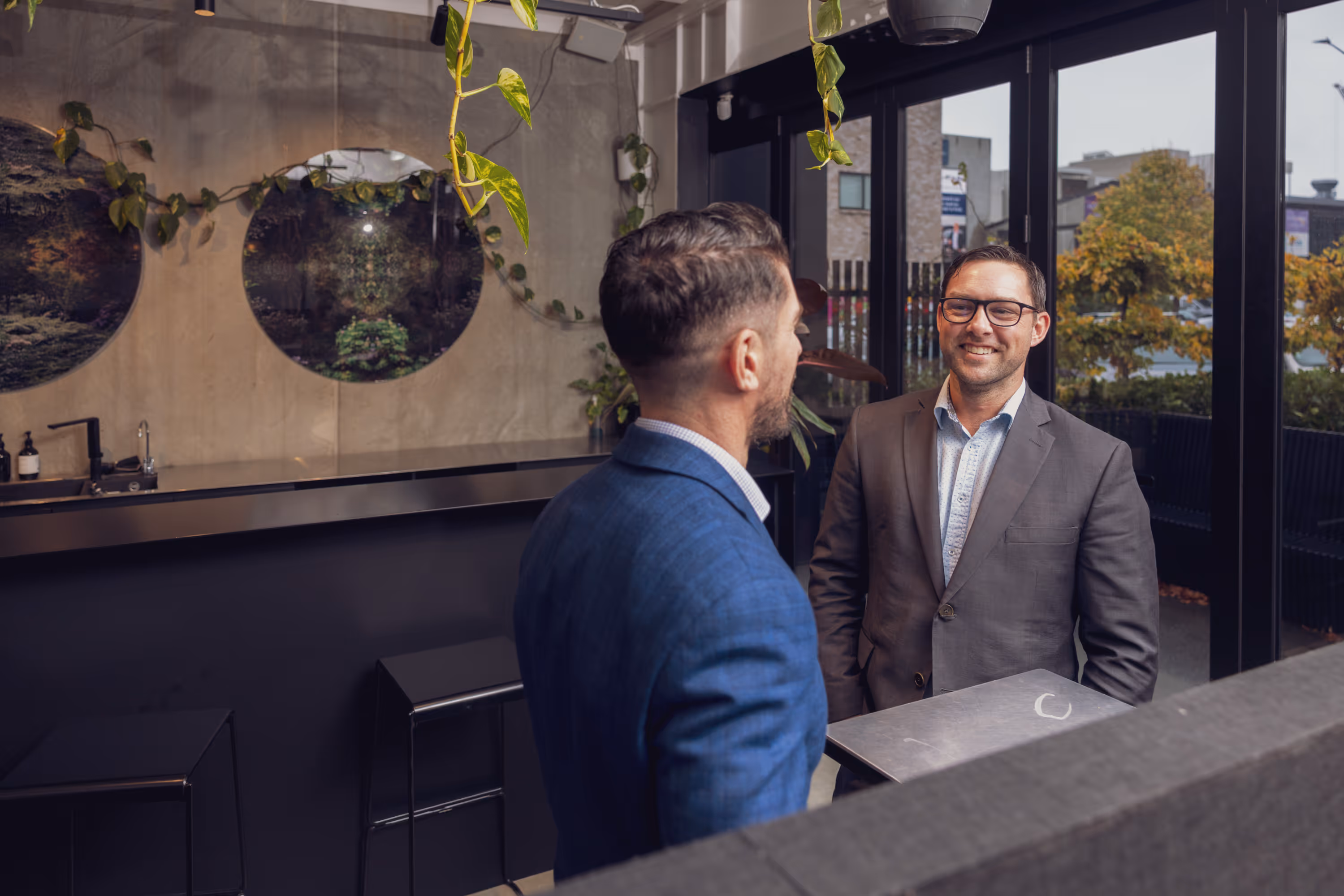 Two men in suits smiling and talking across a counter in a modern office lobby with hanging plants and large windows.
