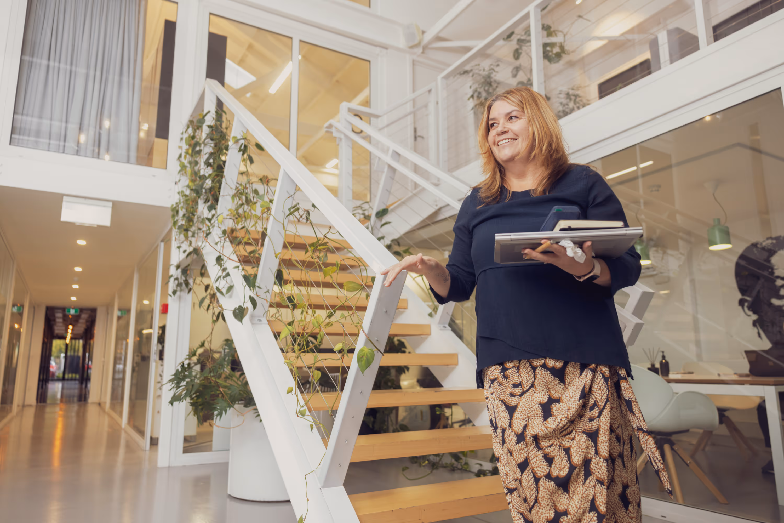 Smiling woman holding notebooks and phone standing near a white staircase with plants in a modern office.