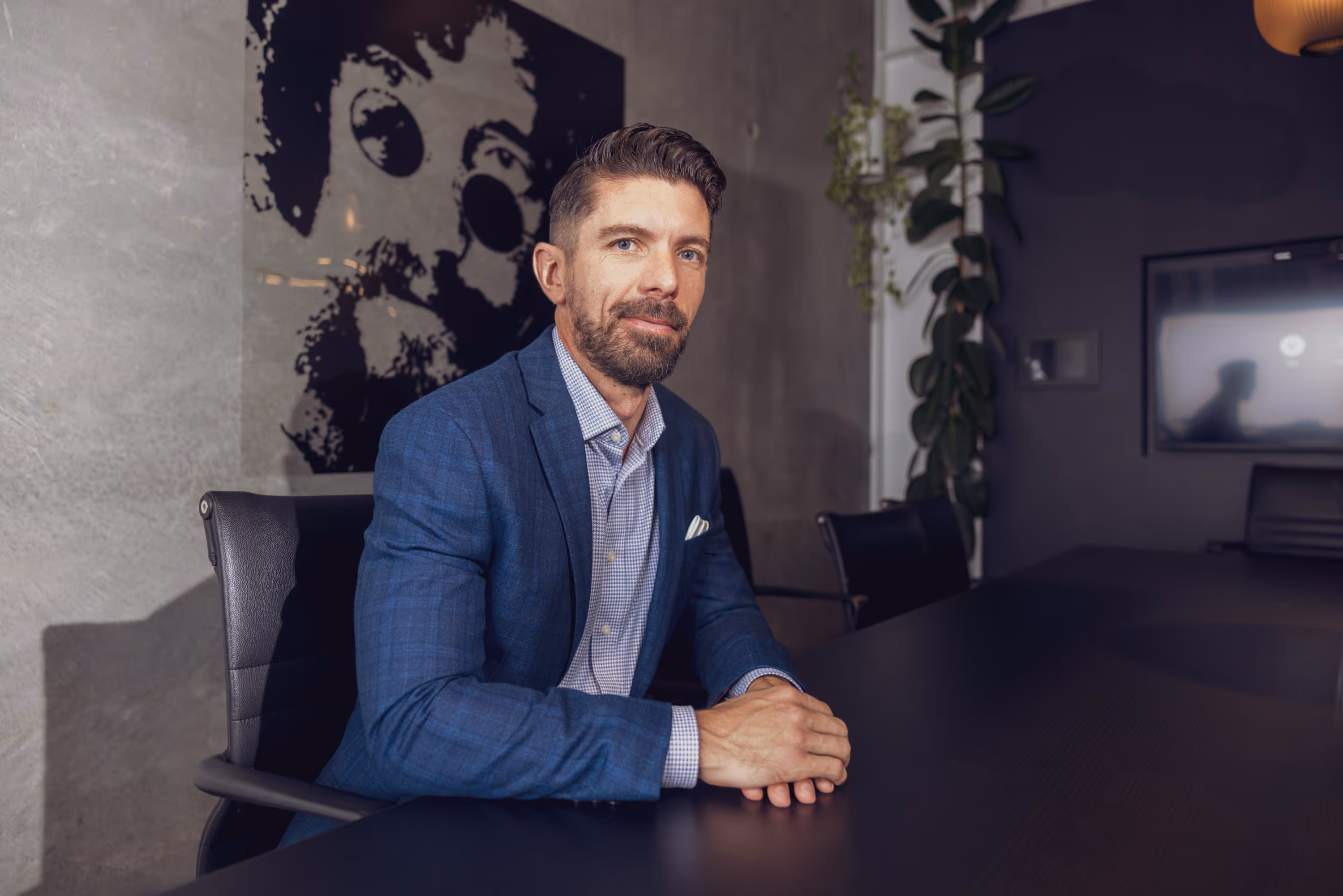 Man in a blue suit sitting at a dark conference table with a modern office background.