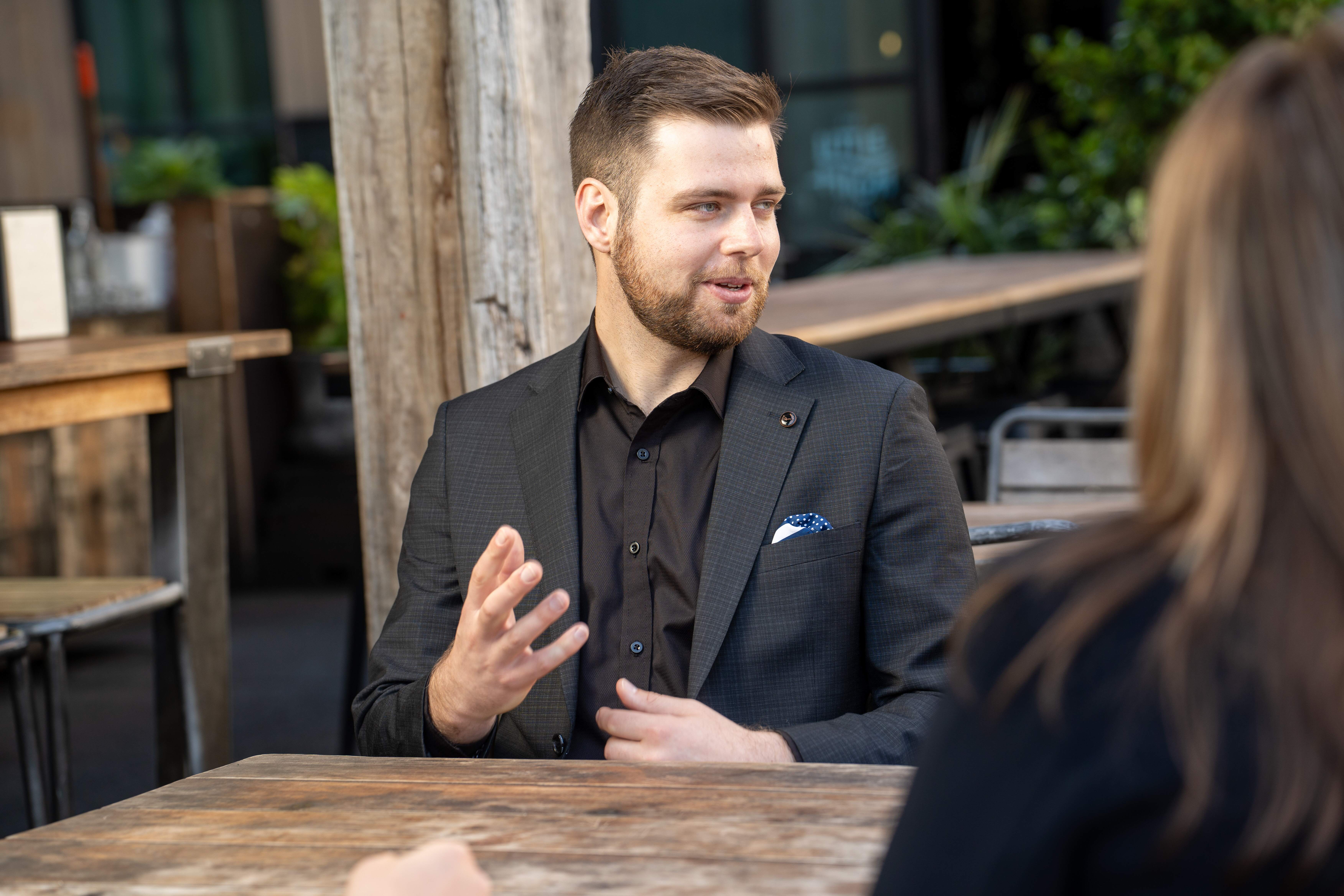 Man in a dark suit gestures while talking to a woman at an outdoor wooden table.