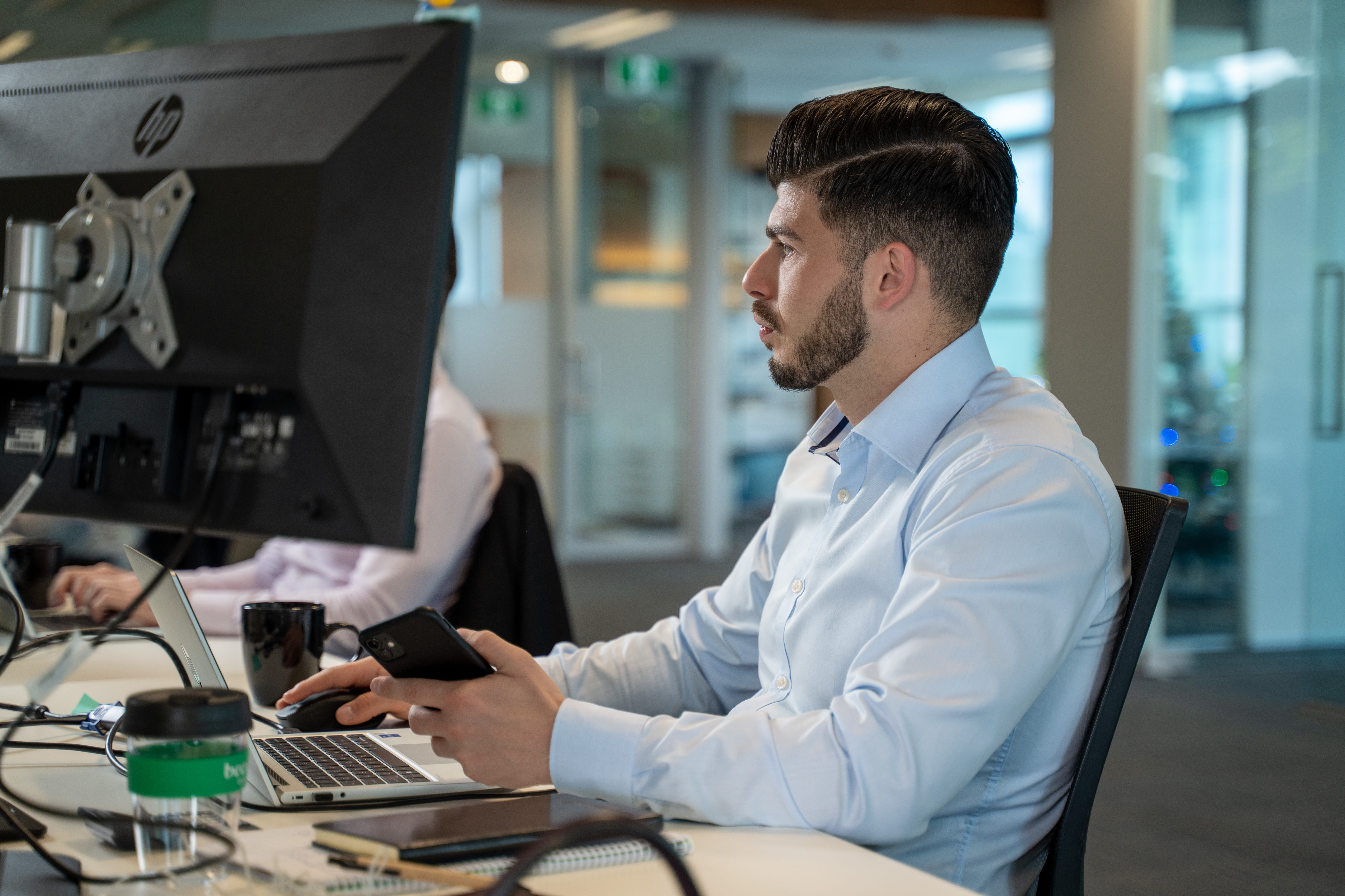 Man with beard sitting at an office desk working on a laptop and holding a smartphone.