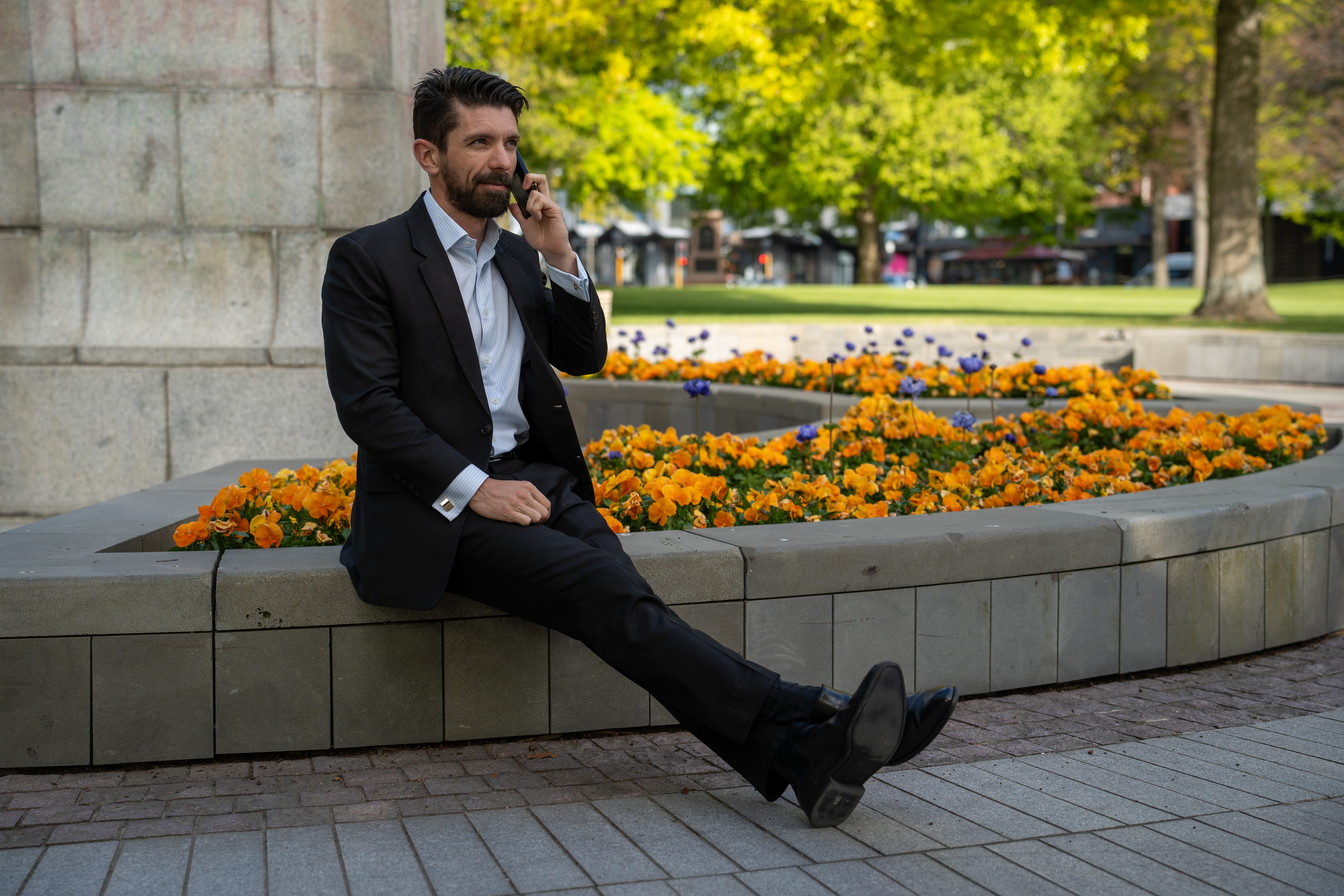 Man in a black suit sitting on a stone ledge in a park, talking on his phone with orange and purple flowers behind him.