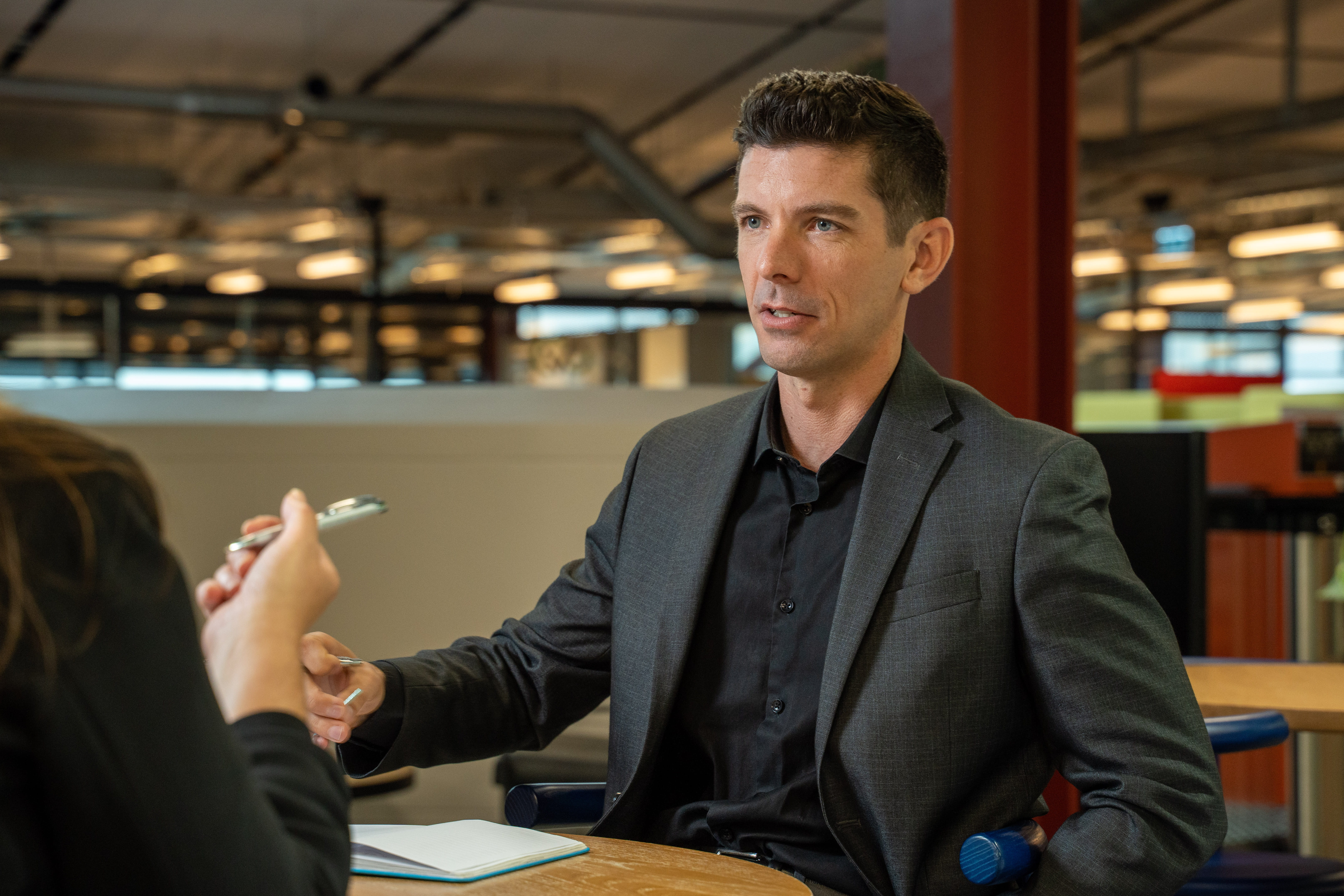 Man in a dark suit and shirt sitting at a table, talking to another person and holding a pen in an office setting.