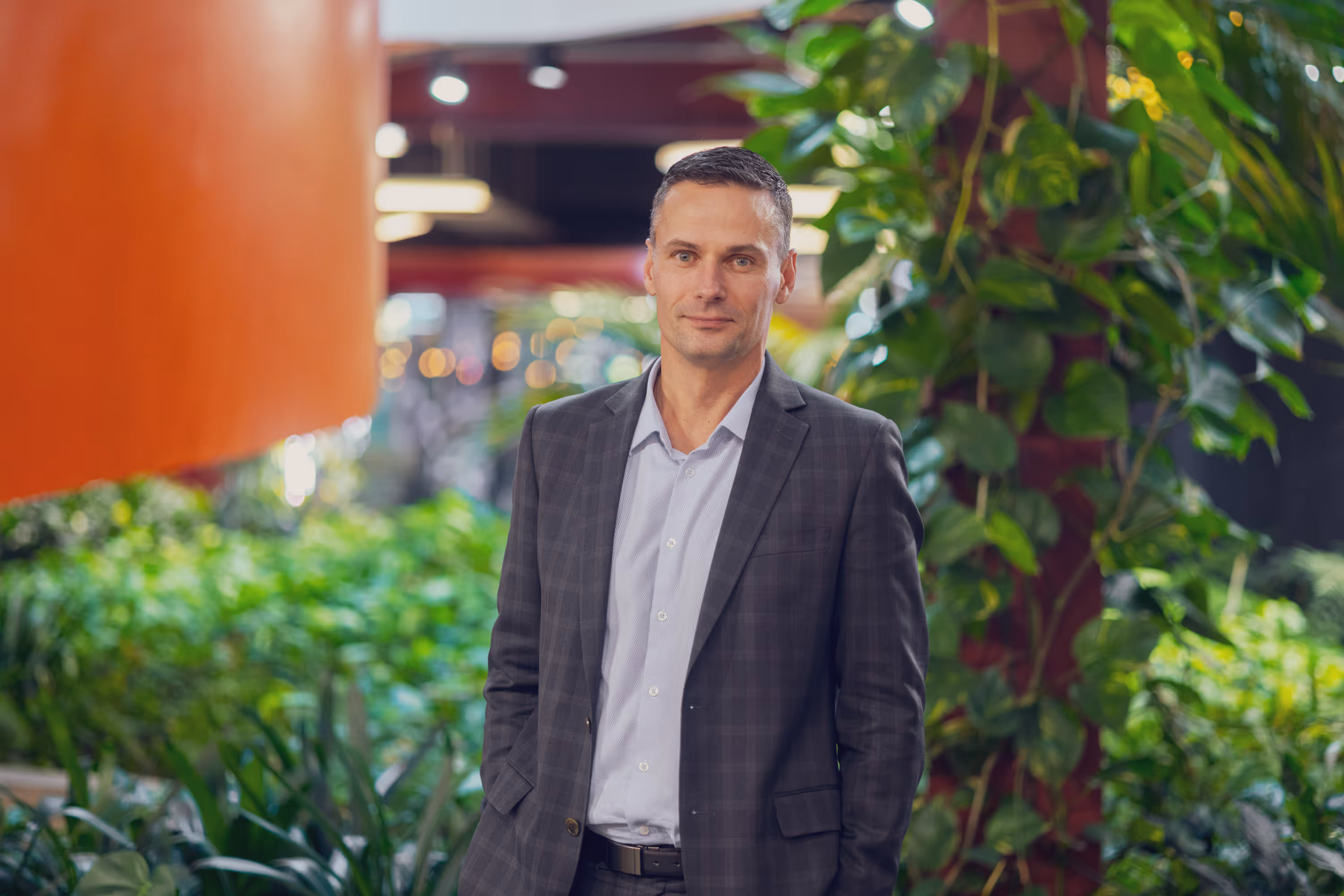 Man in a dark plaid suit standing in an indoor garden with green plants and orange wall behind.