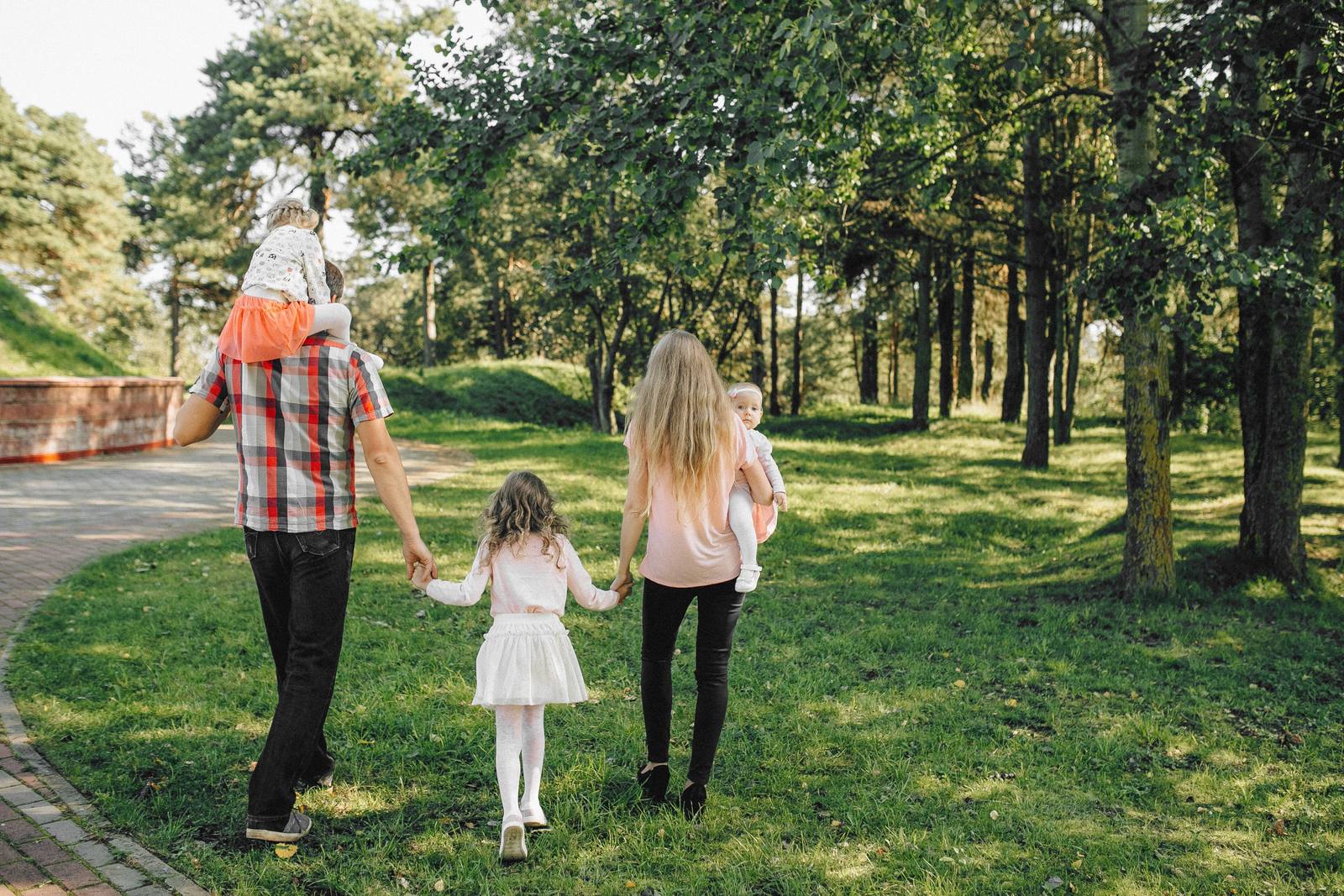 Two parents walking through a sunlit park with three young children, one on his shoulders and one held in her arms