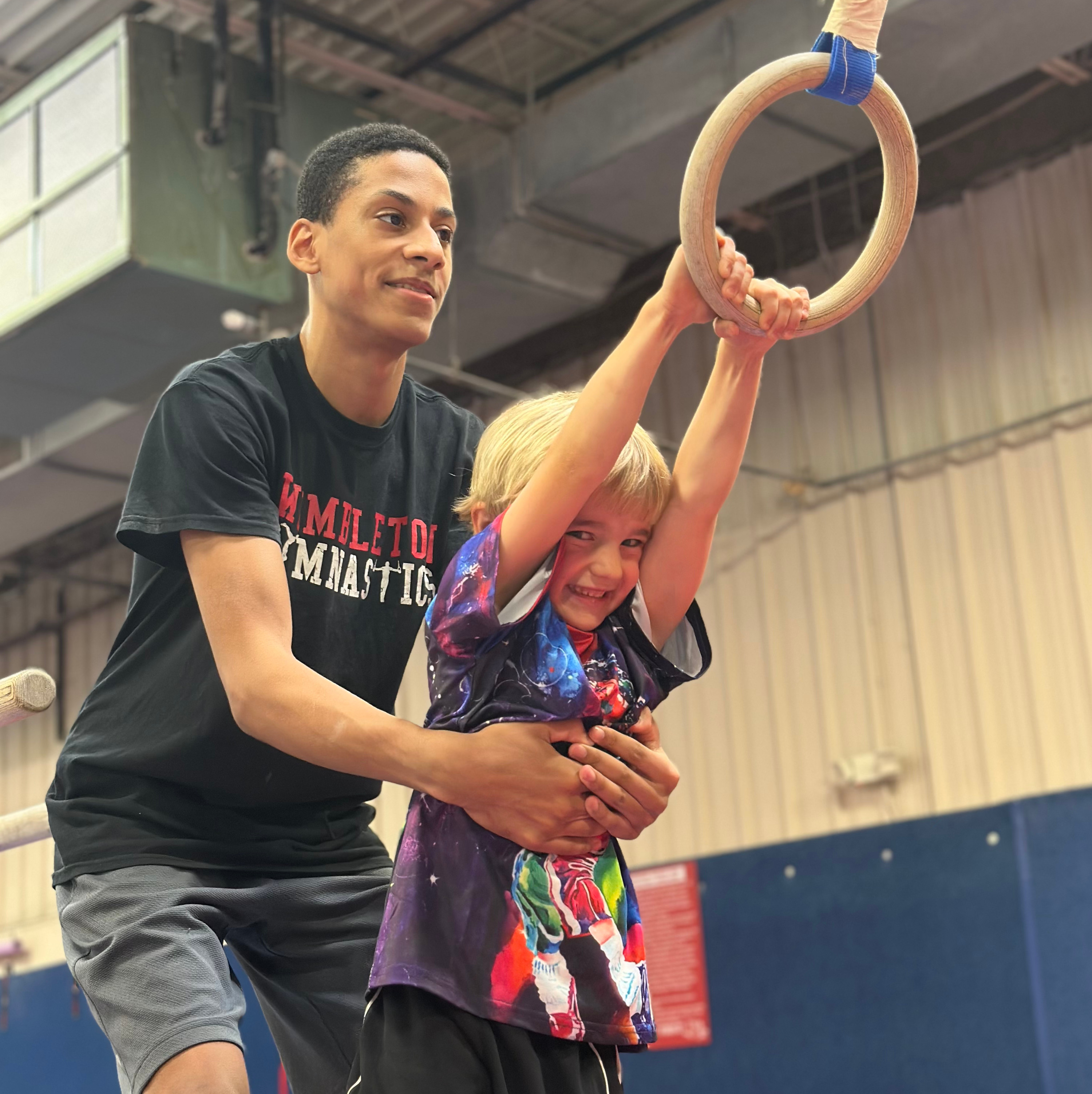 Young male coach helping a smiling boy hold onto wooden gymnastics rings indoors.