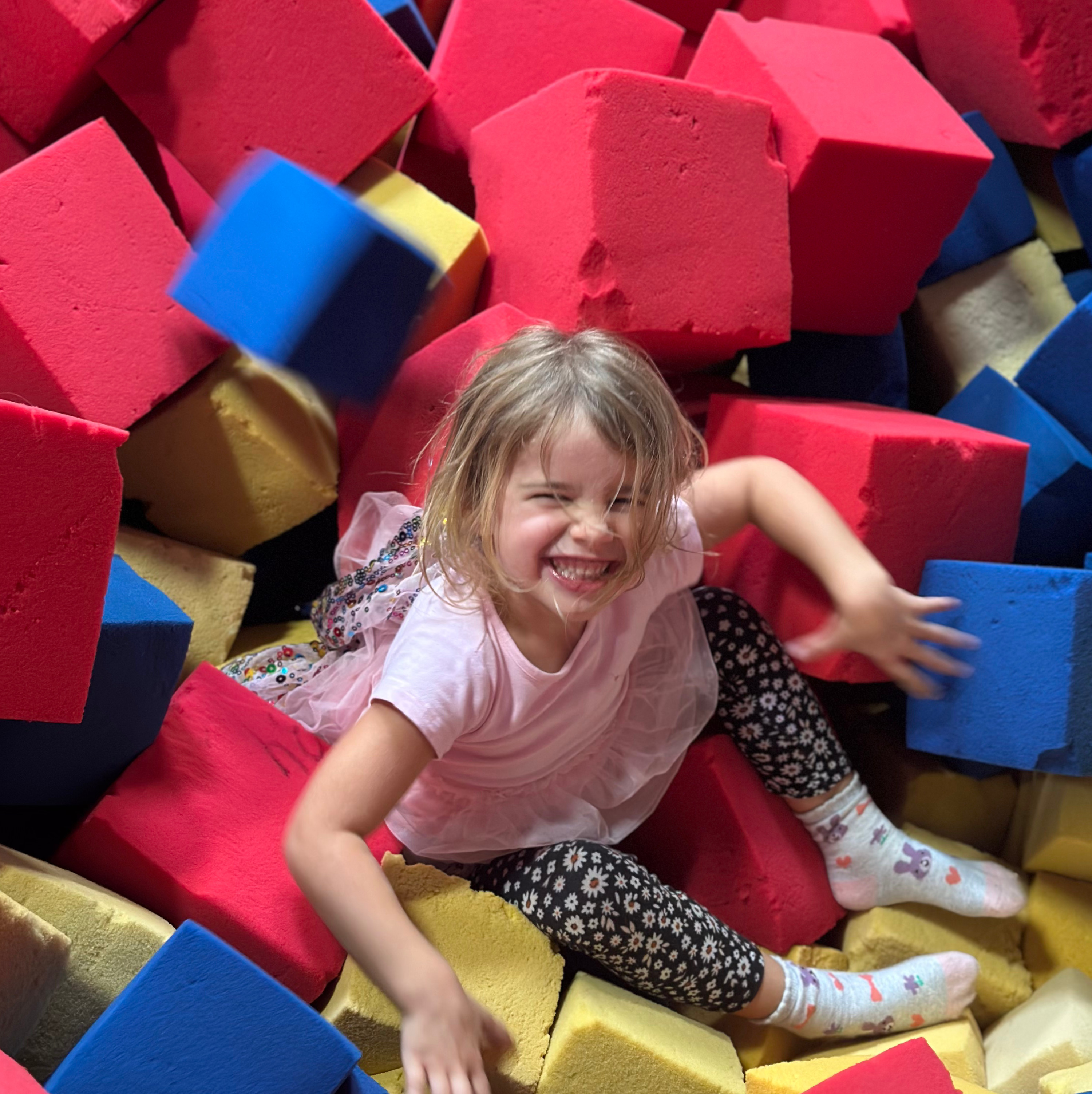 Smiling young girl playing in a pit filled with large red, blue, and yellow foam blocks.