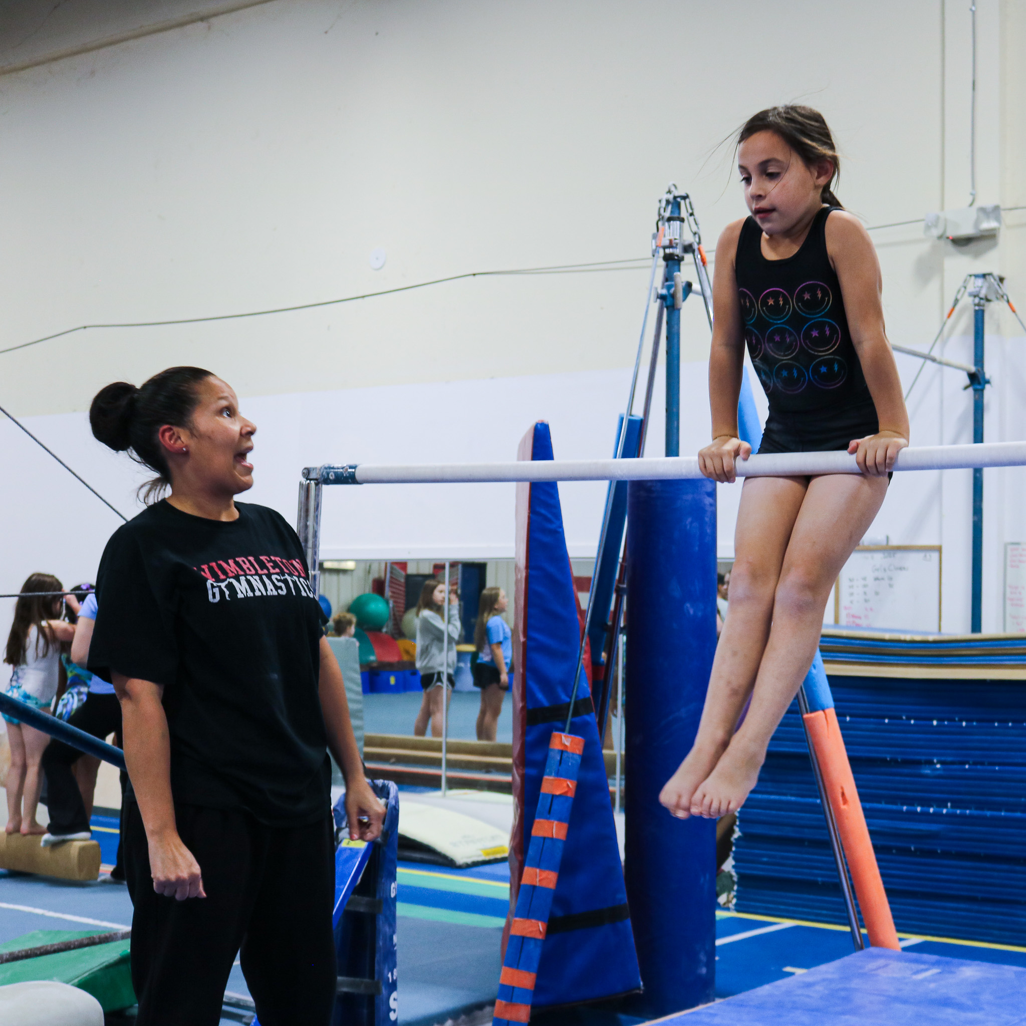 Gymnastics coach instructing a young girl hanging on uneven bars in a gymnasium.