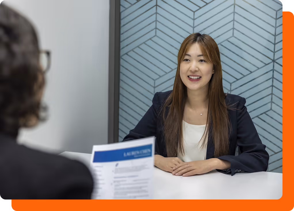 Smiling woman in a blazer sitting at a desk during a job interview with an interviewer holding a resume.